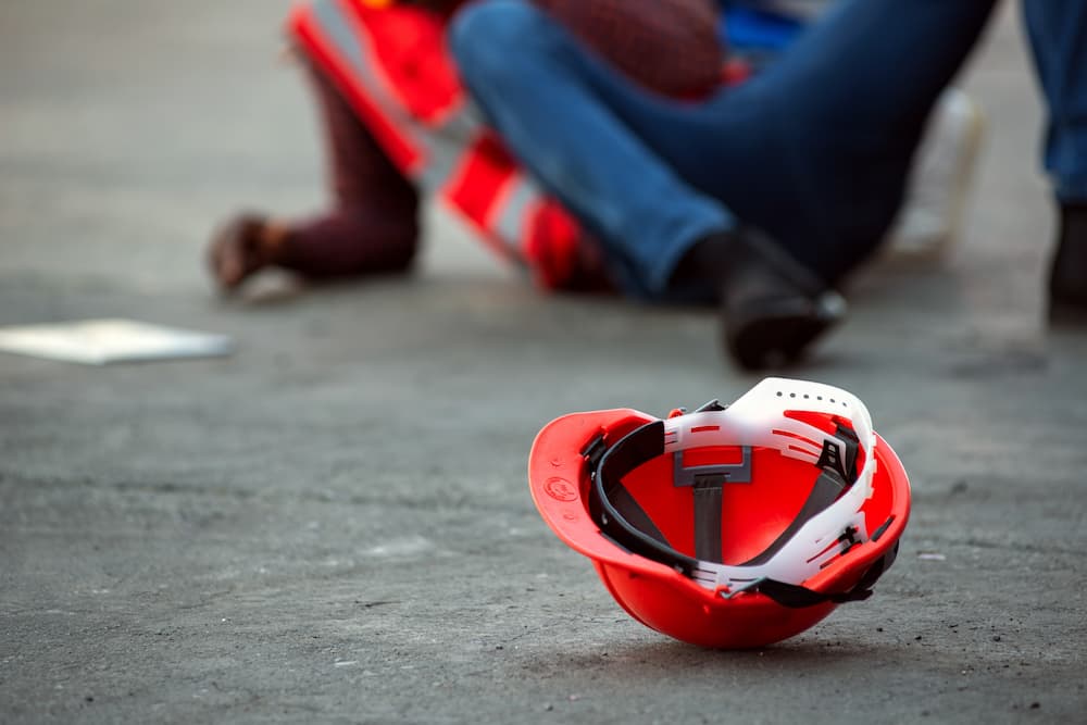 Bright orange hard hat lying on the ground in the foreground, with injured construction workers blurred in the background, symbolizing a construction workplace injury and the need for medical and legal assistance.